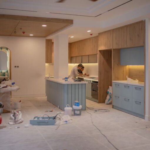Kitchen under renovation with a worker at an island, blue cabinets, and construction tools scattered on the floor.