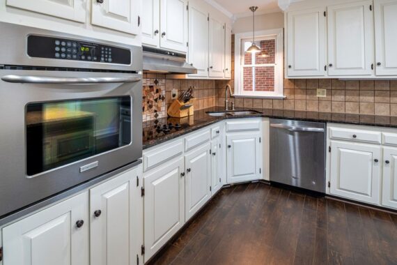 Modern L-shaped kitchen with white cabinets, stainless steel double oven and dishwasher, dark granite counters, and tile backsplash.