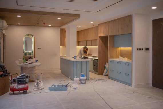 A kitchen under renovation with a worker at a central island, blue base cabinets, wooden upper cabinetry, and construction tools scattered on the floor.